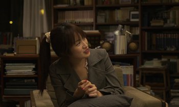 Movie still from “Like Someone in Love” (2012), directed by Abbas Kiarostami – A woman sitting on top of a couch in front of a bookcase; Medium shot, Over the shoulder angle