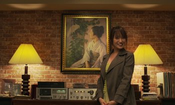 Movie still from “Like Someone in Love” (2012), directed by Abbas Kiarostami – A woman standing in front of a brick wall with a painting on the wall; Medium shot, Low angle
