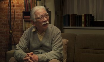 Movie still from “Like Someone in Love” (2012), directed by Abbas Kiarostami – An older man sitting on a couch in a living room; Medium shot, Over the shoulder angle