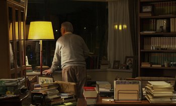 Movie still from “Like Someone in Love” (2012), directed by Abbas Kiarostami – An older man standing in a cluttered living room; Wide shot, Over the shoulder angle