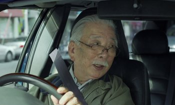Movie still from “Like Someone in Love” (2012), directed by Abbas Kiarostami – An older man driving a car while holding a steering wheel; Close Up shot, Over the shoulder angle
