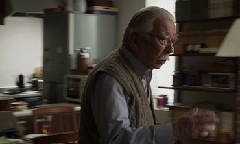 Movie still from “Like Someone in Love” (2012), directed by Abbas Kiarostami – An old man in a sweater is standing in a kitchen; Medium shot, Over the shoulder angle