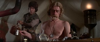 Movie still from “Little Big Man” (1970), directed by Arthur Penn – A man with long blond hair and a beard is sitting in front of another man with long blond hair and a beard; Medium shot, Low angle