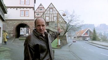 Movie still from “Little Dieter Needs to Fly” (1997), directed by Werner Herzog – A man standing on the side of a road near a building; Medium shot, Low angle