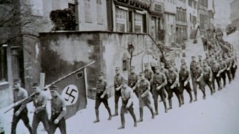 Movie still from “Little Dieter Needs to Fly” (1997), directed by Werner Herzog – A black and white photo of a group of men marching down a street; Wide shot, High angle