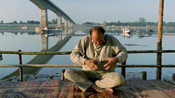Movie still from “Little Dieter Needs to Fly” (1997), directed by Werner Herzog – A man sitting on the ground near a body of water; Wide shot, Low angle
