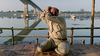 Movie still from “Little Dieter Needs to Fly” (1997), directed by Werner Herzog – A man sitting on the ground in front of a body of water; Medium shot, Low angle