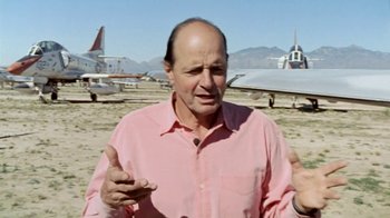 Movie still from “Little Dieter Needs to Fly” (1997), directed by Werner Herzog – A man in a pink shirt is standing in front of some airplanes; Medium shot, Low angle