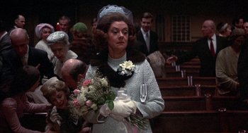 Movie still from “Little Murders” (1971), directed by Alan Arkin – An older woman holding a bouquet of flowers in a church; Medium shot, Over the shoulder angle