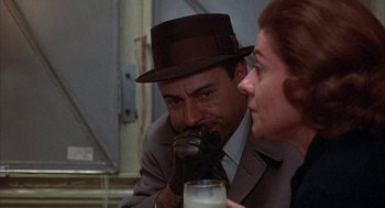 Movie still from “Little Murders” (1971), directed by Alan Arkin – A man in a suit and a woman in a brown hat are looking at a glass; Close Up shot, Over the shoulder angle