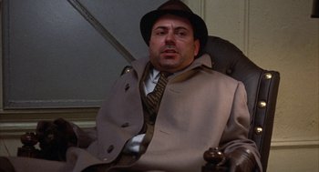 Movie still from “Little Murders” (1971), directed by Alan Arkin – A man wearing a hat and a coat sitting in a chair; Medium shot, Low angle