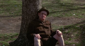 Movie still from “Little Murders” (1971), directed by Alan Arkin – An older man sitting on the ground next to a tree; Medium shot, High angle