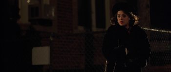 Movie still from “Little Odessa” (1994), directed by James Gray – A woman in a black coat and hat standing in front of a chain link fence at night; Medium shot, Over the shoulder angle