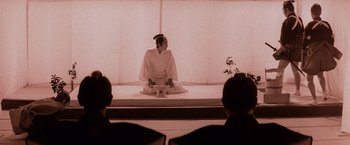 Movie still from “Lone Wolf and Cub: Baby Cart to Hades” (1972), directed by Kenji Misumi – A person sitting on a stage in front of a crowd; Wide shot, Low angle