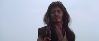 Movie still from “Lone Wolf and Cub: Baby Cart to Hades” (1972), directed by Kenji Misumi – A man with long hair holding a gun in his hand; Close Up shot, Low angle