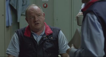 Movie still from “Looking for Eric” (2009), directed by Ken Loach – A man wearing a black vest is talking to someone; Medium shot, Over the shoulder angle