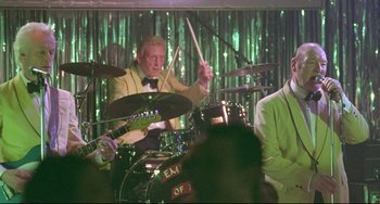 Movie still from “Looking for Eric” (2009), directed by Ken Loach – An older man playing drums in front of a group of people; Medium shot, Over the shoulder angle