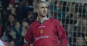 Movie still from “Looking for Eric” (2009), directed by Ken Loach – A man in a red soccer uniform stands in front of a crowd of people; Close Up shot, High angle