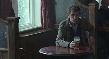Movie still from “Looking for Eric” (2009), directed by Ken Loach – A man sitting at a table holding a glass of beer; Medium shot, High angle