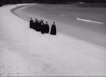 Movie still from “Lord of the Flies” (1963), directed by Peter Brook – A black and white photo of a group of people in black robes on the beach; Extreme Wide shot, High angle