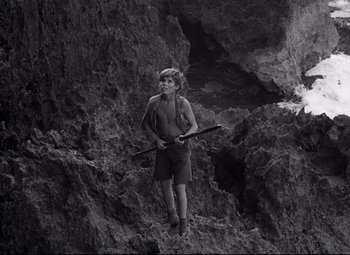 Movie still from “Lord of the Flies” (1963), directed by Peter Brook – A young man holding a rifle standing on a rock; Wide shot, High angle