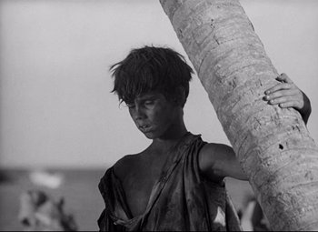 Movie still from “Lord of the Flies” (1963), directed by Peter Brook – A young man standing next to a palm tree; Medium shot, Low angle