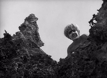 Movie still from “Lord of the Flies” (1963), directed by Peter Brook – A man with blonde hair standing next to some rocks; Medium shot, Low angle