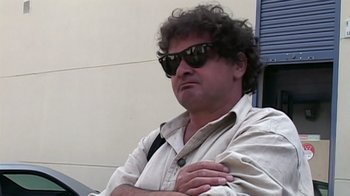 Movie still from “Lost in La Mancha” (2002), directed by Louis Pepe – A man wearing sunglasses sitting in front of a building; Close Up shot, Low angle