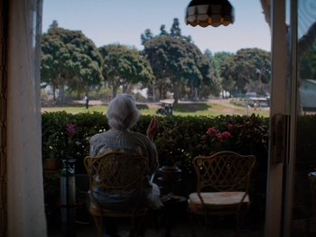 Movie still from “Lucy in the Sky” (2019), directed by Noah Hawley – An older woman sitting on a chair looking out a window at a park; Wide shot, Over the shoulder angle