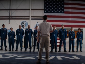 Movie still from “Lucy in the Sky” (2019), directed by Noah Hawley – A group of men in blue suits standing in front of an american flag; Wide shot, Over the shoulder angle