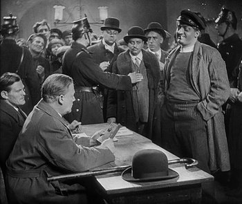 Movie still from “M” (1931), directed by Fritz Lang – A group of men standing around a table with a man sitting at a desk; Medium shot, Over the shoulder angle