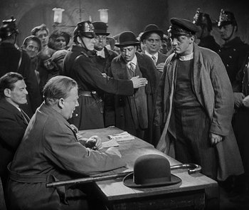Movie still from “M” (1931), directed by Fritz Lang – A group of men standing around a table with a man sitting at a table; Medium shot, Over the shoulder angle