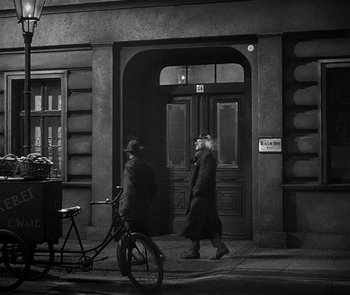 Movie still from “M” (1931), directed by Fritz Lang – Two people walking down a street near a bicycle; Wide shot, Over the shoulder angle