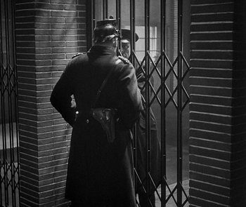 Movie still from “M” (1931), directed by Fritz Lang – A man in a black coat standing next to a gate; Medium shot, Over the shoulder angle