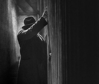 Movie still from “M” (1931), directed by Fritz Lang – A man in a hat and a long coat leaning against a wall; Medium shot, Low angle
