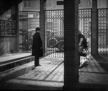 Movie still from “M” (1931), directed by Fritz Lang – A man in a black coat and a black hat standing in front of a gate; Extreme Wide shot, High angle
