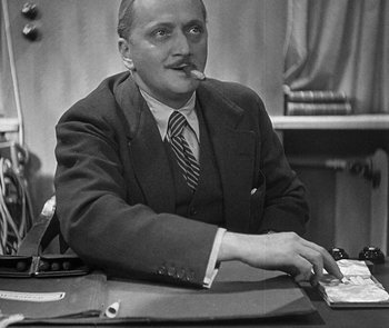 Movie still from “M” (1931), directed by Fritz Lang – An older man sitting at a desk with a cigarette in his mouth; Medium shot, Low angle