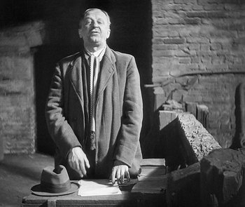 Movie still from “M” (1931), directed by Fritz Lang – An older man in a suit and tie sitting on top of a suitcase; Medium shot, Low angle