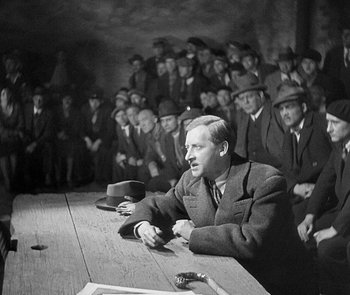 Movie still from “M” (1931), directed by Fritz Lang – A man sitting at a table in front of a group of men; Medium shot, High angle