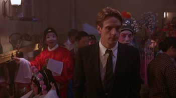 Movie still from “M. Butterfly” (1993), directed by David Cronenberg – A man wearing a suit and tie standing next to a crowd of people; Close Up shot, Over the shoulder angle