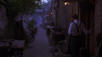 Movie still from “M. Butterfly” (1993), directed by David Cronenberg – A man standing on a sidewalk near a building; Wide shot, Over the shoulder angle
