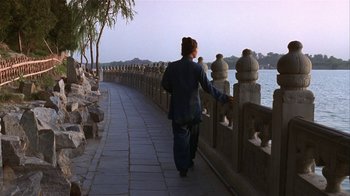 Movie still from “M. Butterfly” (1993), directed by David Cronenberg – A person walking on a sidewalk near a body of water; Wide shot, Over the shoulder angle