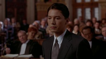Movie still from “M. Butterfly” (1993), directed by David Cronenberg – A man wearing a suit and tie in front of a group of people; Close Up shot, High angle