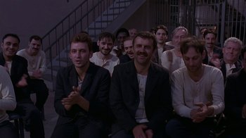 Movie still from “M. Butterfly” (1993), directed by David Cronenberg – A group of men sitting next to each other in front of a staircase; Wide shot, High angle