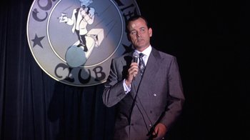 Movie still from “Mad Dog and Glory” (1993), directed by John McNaughton – A man in a suit and tie holding a microphone; Medium shot, Low angle