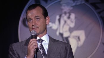 Movie still from “Mad Dog and Glory” (1993), directed by John McNaughton – A man wearing a suit and tie holding a microphone; Close Up shot, Over the shoulder angle