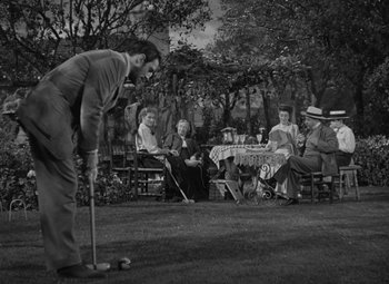 Movie still from “Madame Curie” (1943), directed by Mervyn LeRoy – A group of people sitting at a table and a man holding a golf club; Wide shot, High angle