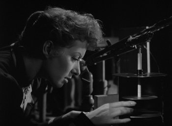 Movie still from “Madame Curie” (1943), directed by Mervyn LeRoy – A woman looking through a microscope in a room; Medium shot, Low angle