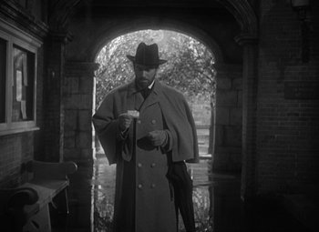 Movie still from “Madame Curie” (1943), directed by Mervyn LeRoy – A black and white photo of a man in a trench coat and a hat; Medium shot, Over the shoulder angle