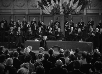 Movie still from “Madame Curie” (1943), directed by Mervyn LeRoy – An old photo of a woman giving a speech in front of an audience; Wide shot, Over the shoulder angle